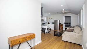 Una sala moderna con un sofá, una mesa de centro y una mesita auxiliar de madera. Se ve una cocina con armarios blancos y electrodomésticos de acero inoxidable al fondo.