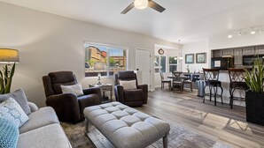 Ein geräumiges Wohnzimmer mit einer Couch, Sesseln und einem Couchtisch. Es gibt einen Essbereich mit Tisch und Stühlen sowie eine Küche mit Barhocker.