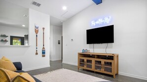 Un salon moderne avec un téléviseur à écran plat fixé au mur, un meuble en bois en dessous, et un coin salon confortable avec un canapé et des coussins.