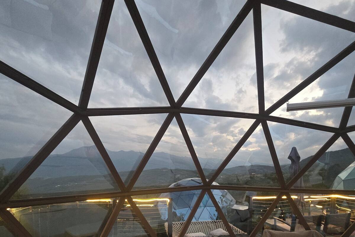 Una habitación con cama, una mesita y una silla, que ofrece vistas a un paisaje urbano y montañas a través de una estructura de vidrio geométrica.