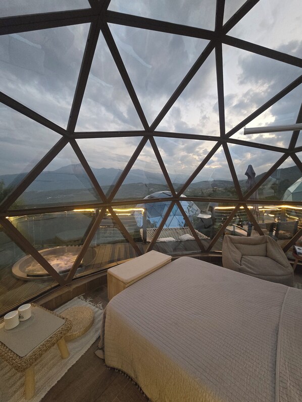 Une pièce comprenant un lit, une petite table et une chaise, avec vue sur un paysage urbain et des montagnes, le tout visible à travers une structure de verre aux formes géométriques.