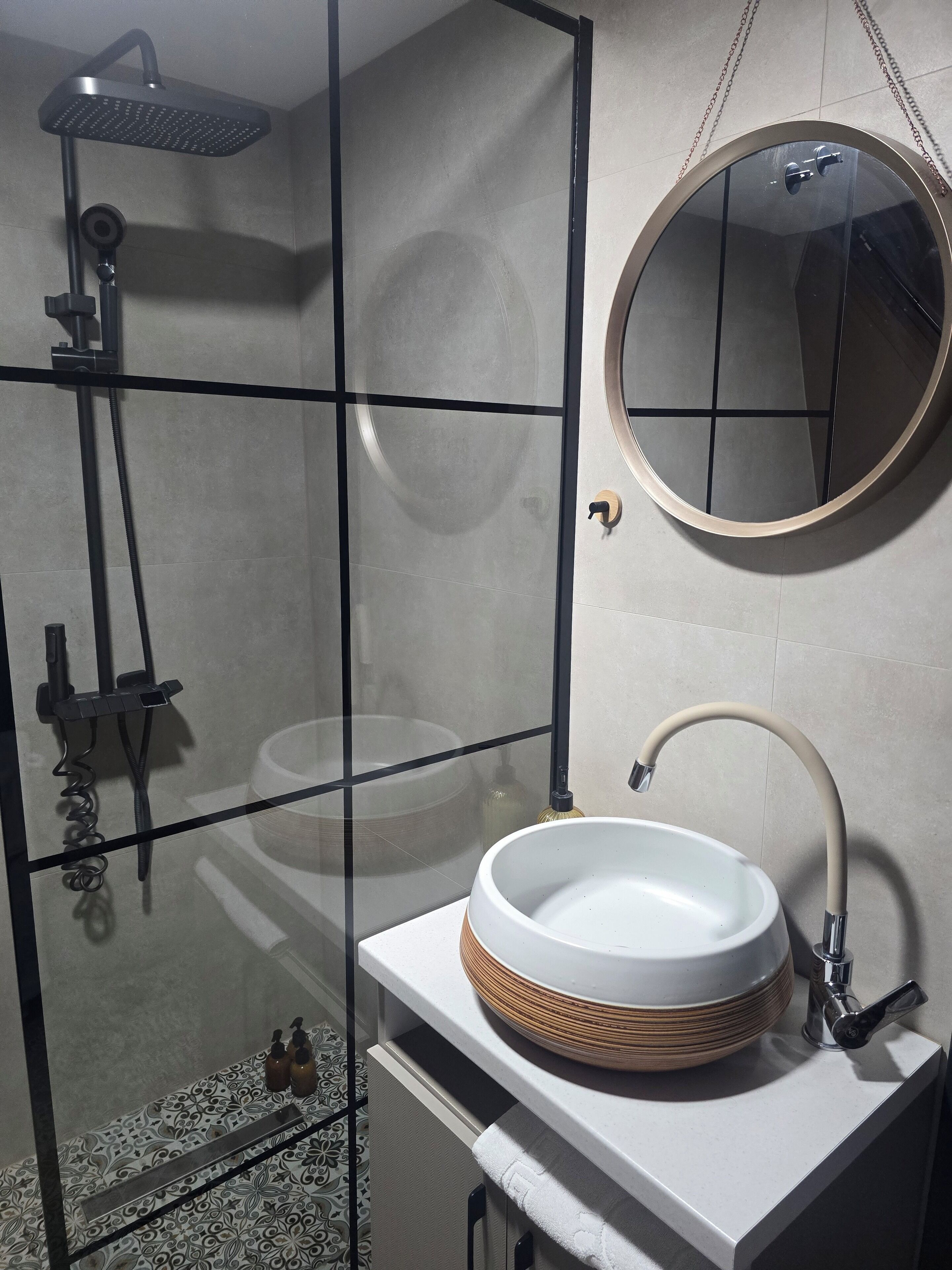 A modern bathroom with a glass-enclosed shower, a round mirror, and a white sink with a beige basin.