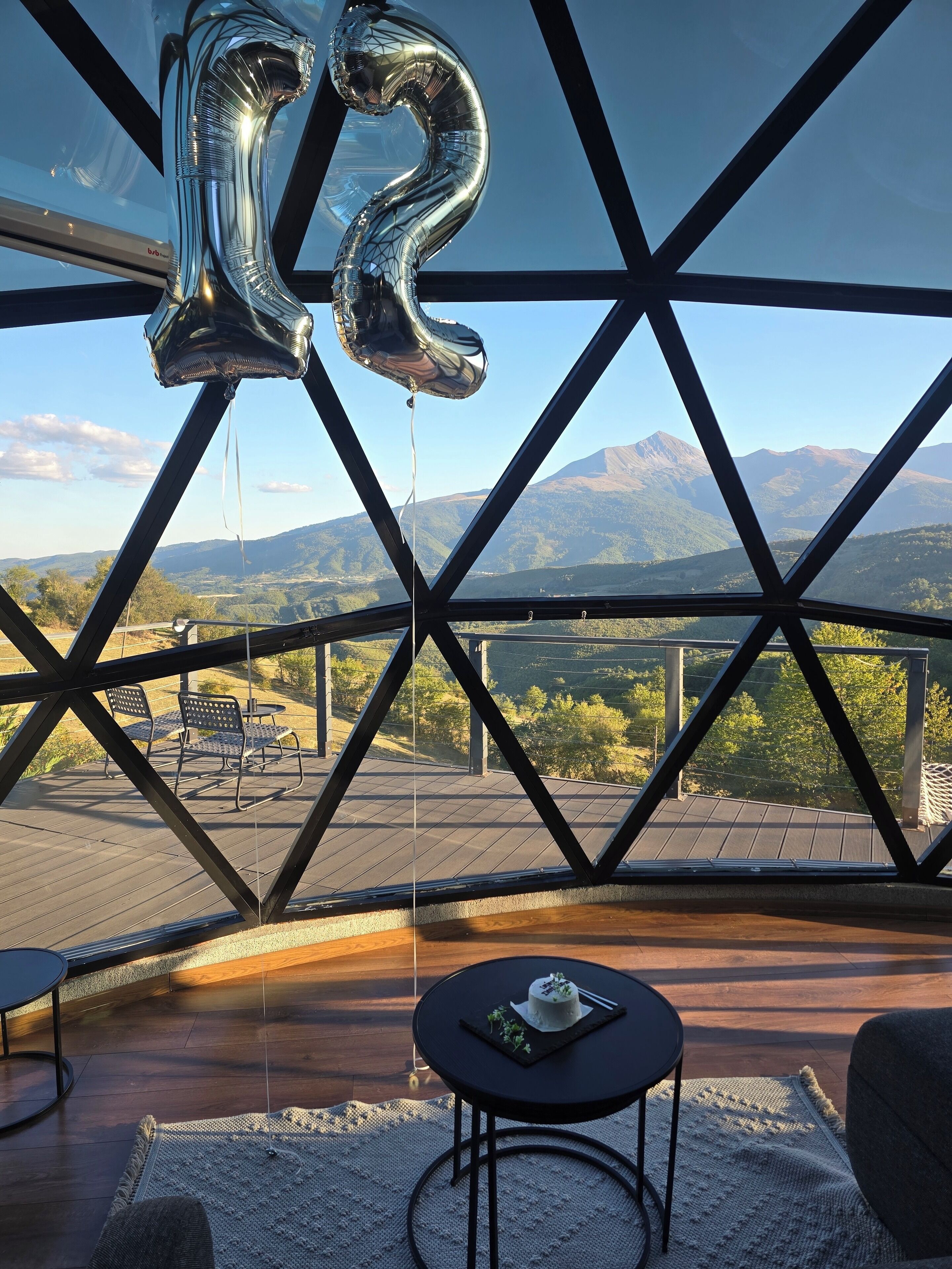 A room with a large window, a metal frame, a table with a chair, and a view of mountains.