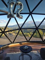 Una habitación con un ventanal grande, estructura metálica, una mesa con una silla y vistas a las montañas.