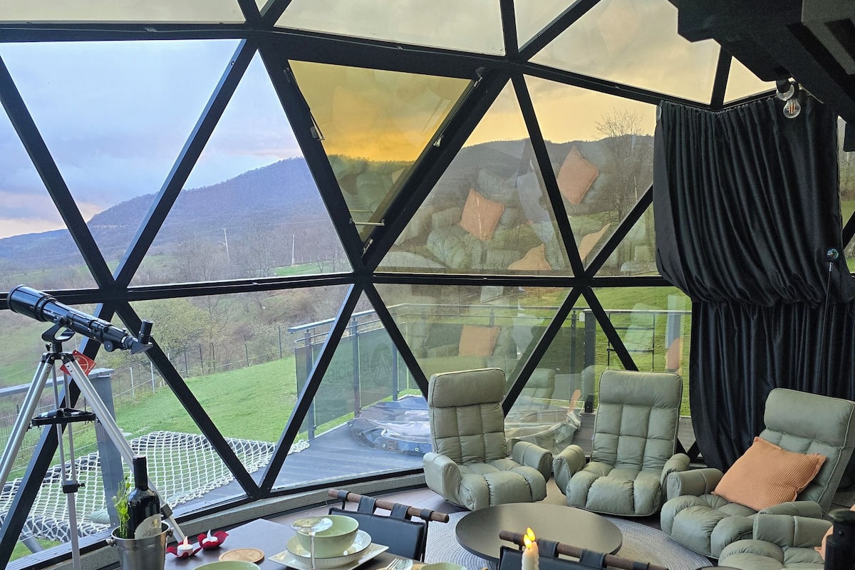 Un comedor con una mesa preparada para dos, un telescopio y una vista panorámica a través de un ventanal grande.