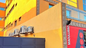 A building with a yellow facade, a red and grey sign reading "MACULUS," and a closed grey gate. - Cozy Modern Studio in Maculusso, Luanda – Central & Secure Location (Luanda)