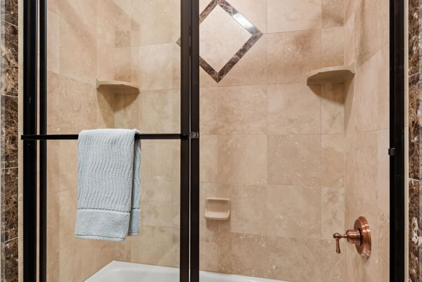 Eine Dusche mit Glastür, beigen Fliesen und einem Handtuch auf einem Haken.