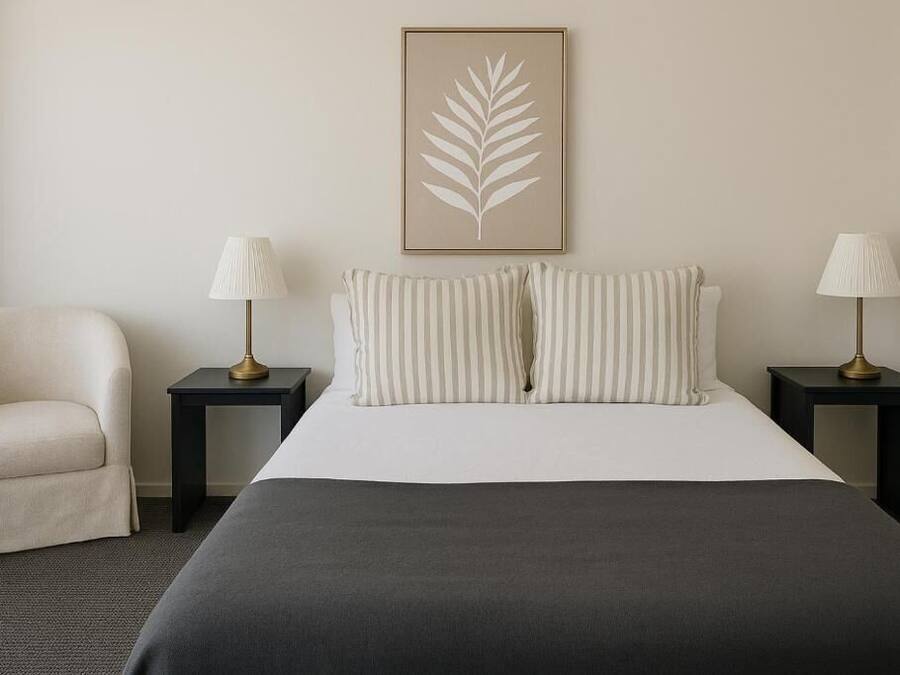 Habitación de hotel con una cama, dos mesitas de noche con lámparas, una silla y un cuadro con hojas en la pared.
