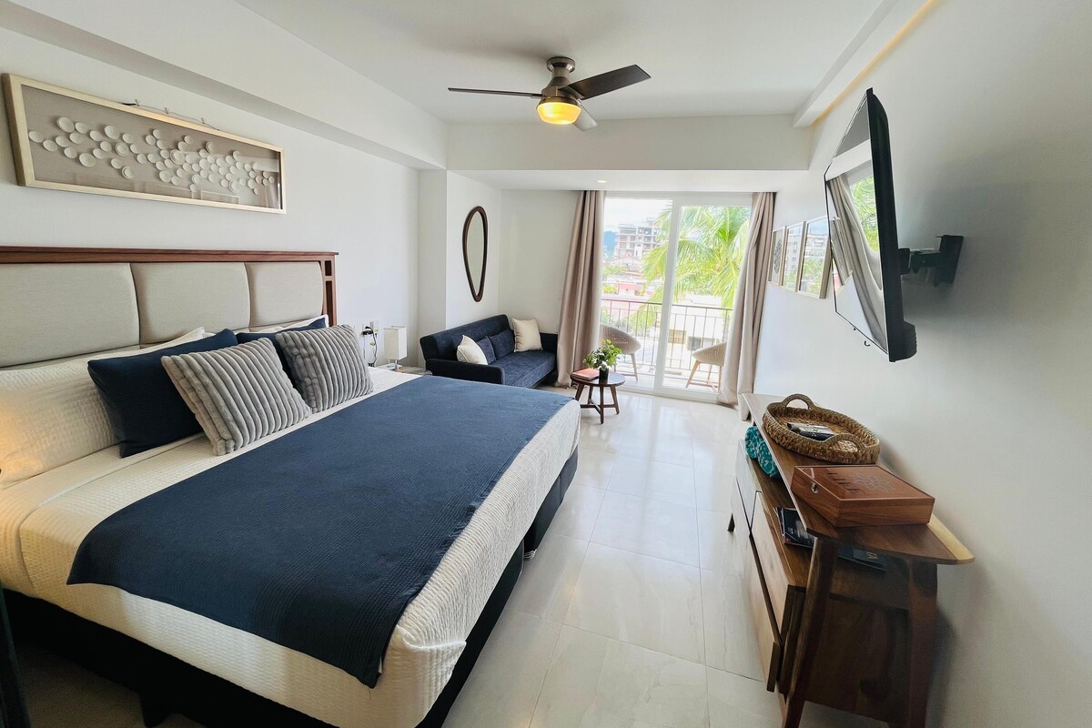 Una habitación de hotel moderna con una cama grande, un ventilador de techo, un televisor montado en la pared y un balcón con vistas a la ciudad.