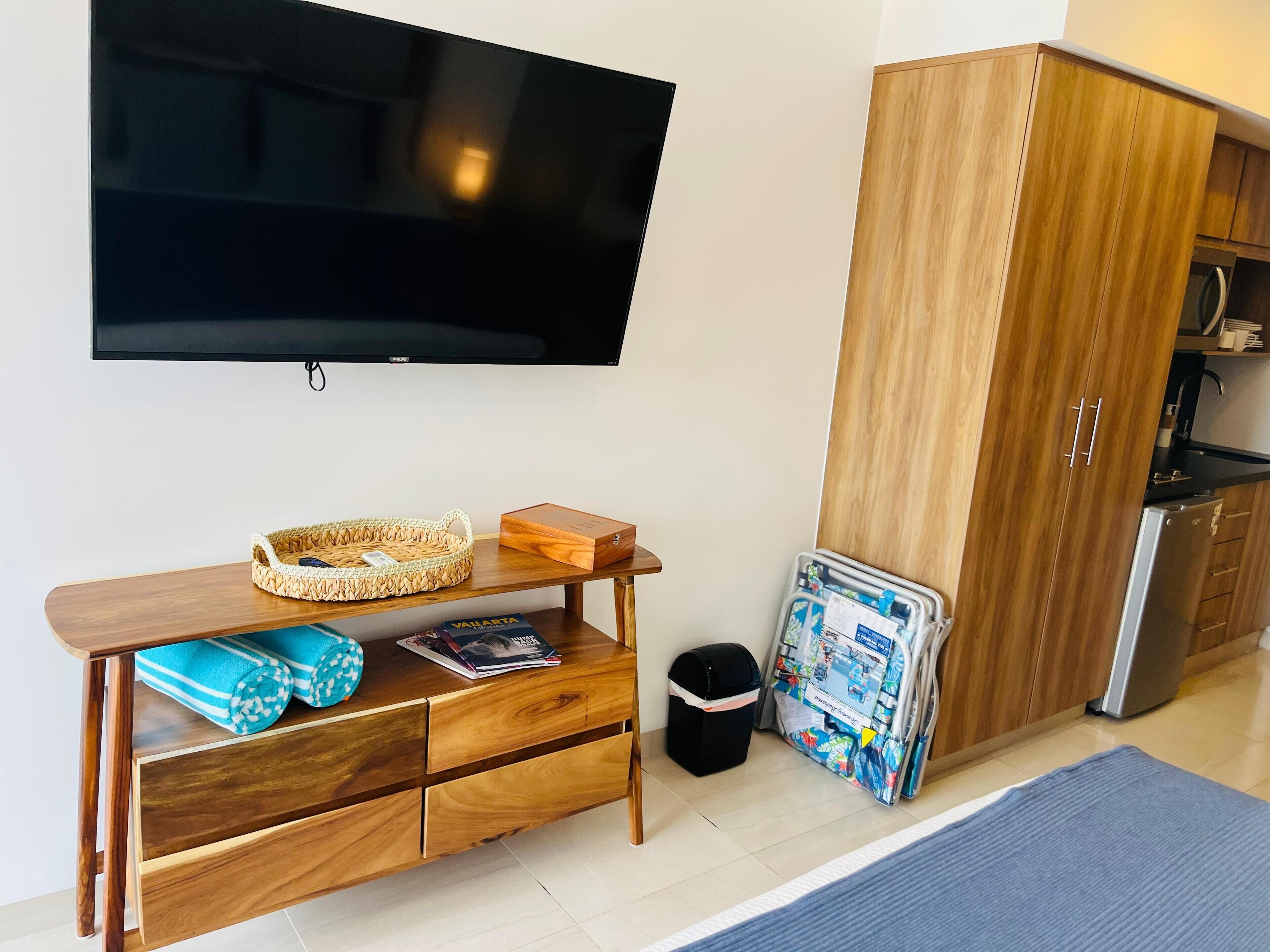 A modern living space with a flat-screen TV, wooden furniture, a kitchen area, and a trash bin.