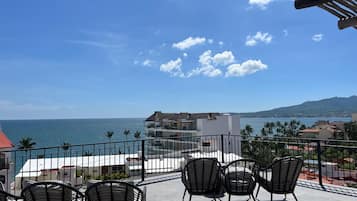 Un balcon avec des chaises donnant sur une vue côtière, comprenant une étendue d’eau et des bâtiments visibles au loin.