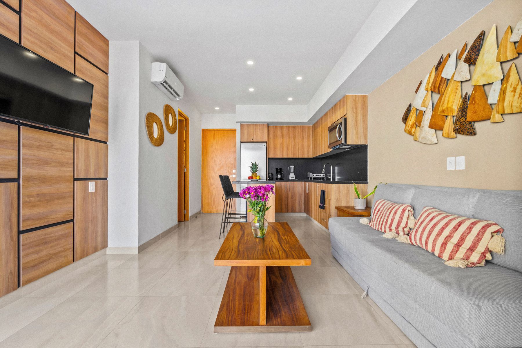 Una cocina moderna con armarios de madera, un comedor con mesa de madera y un sofá gris con cojines a rayas.