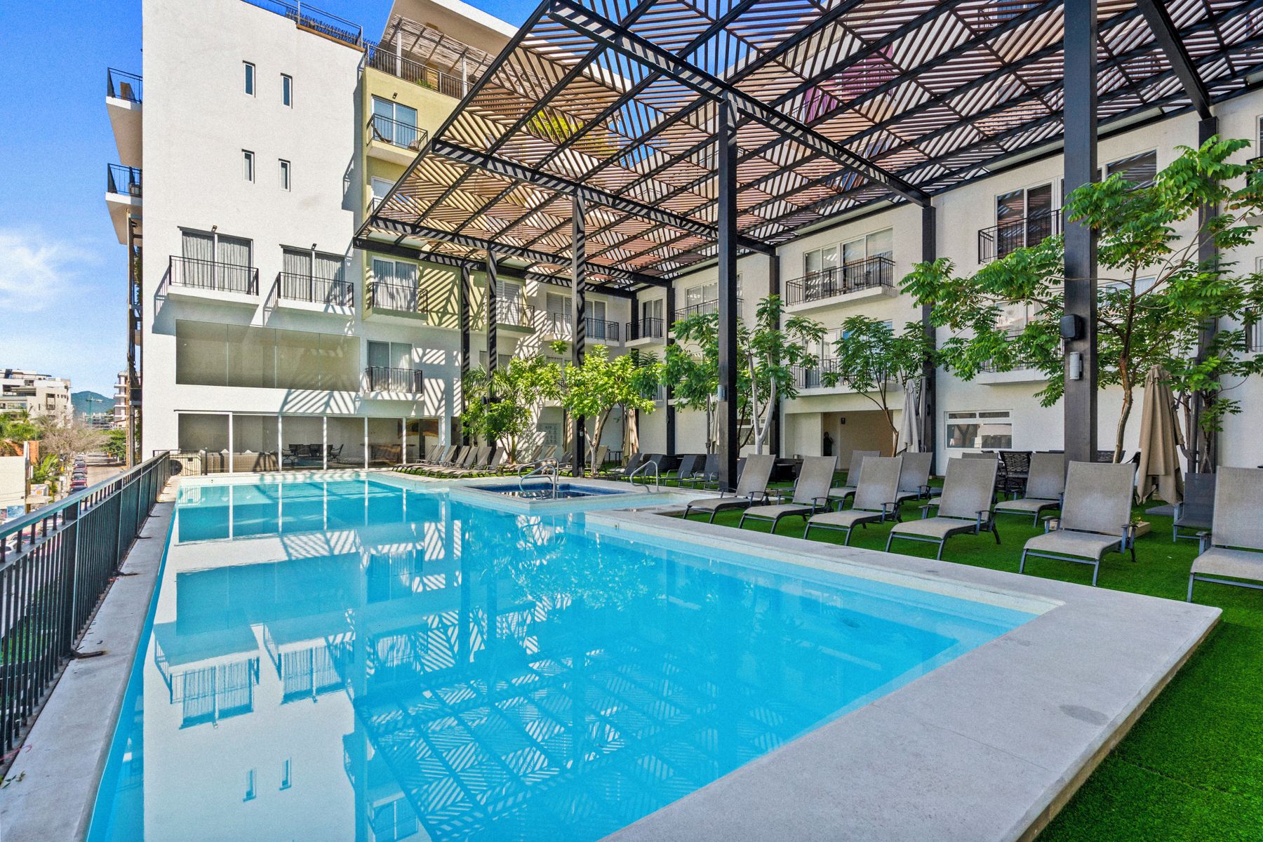 Una piscina con techo de estructura metálica, rodeada de sillas de descanso y follaje.