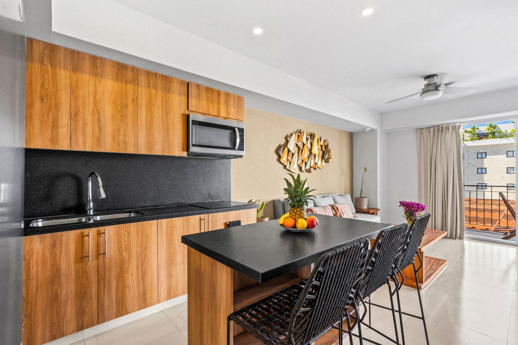 Una cocina moderna con armarios de madera, encimera negra y microondas. Hay una zona de comedor con una mesa y sillas negras, y un frutero sobre la mesa.