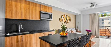 Una cocina moderna con armarios de madera, encimera negra y microondas. Hay una zona de comedor con una mesa y sillas negras, y un frutero sobre la mesa.