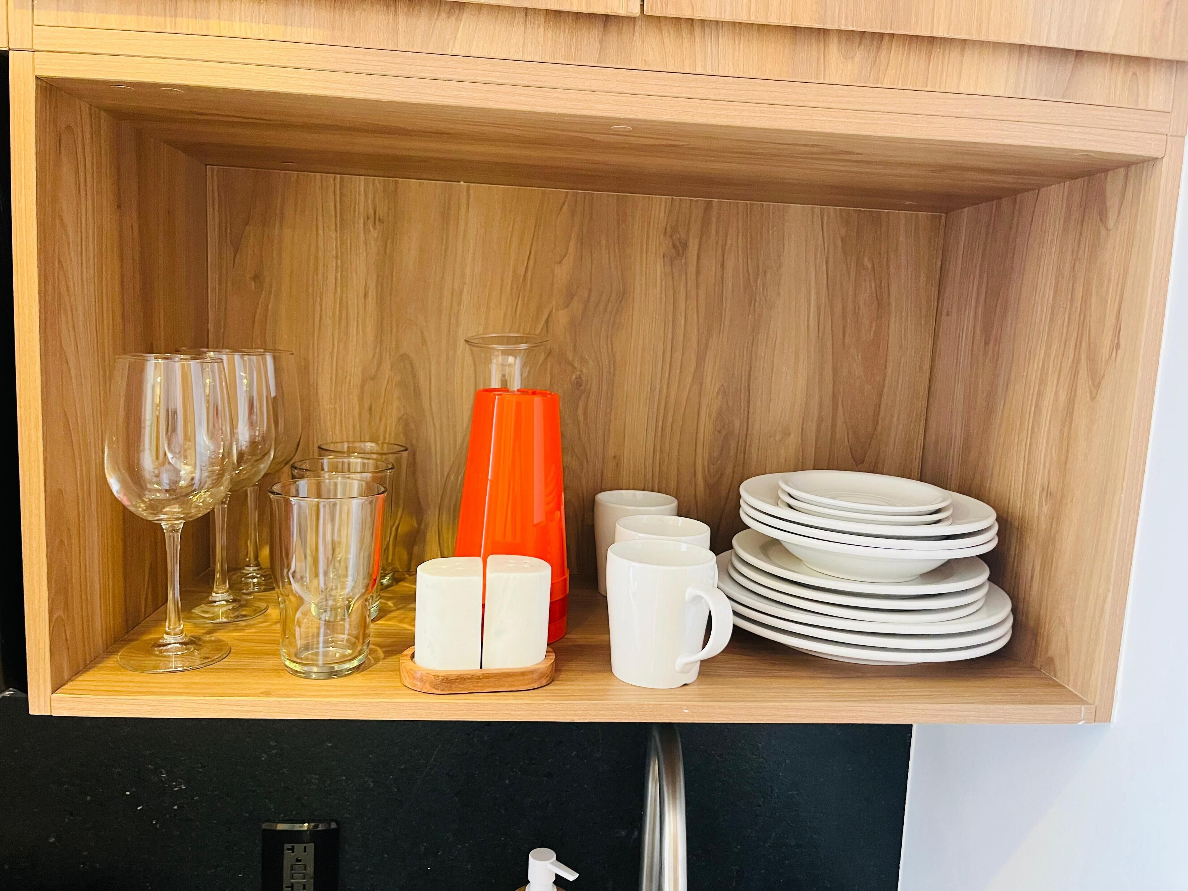 A wooden cabinet with wine glasses, a glass carafe, a red vase, and stacked white plates.
