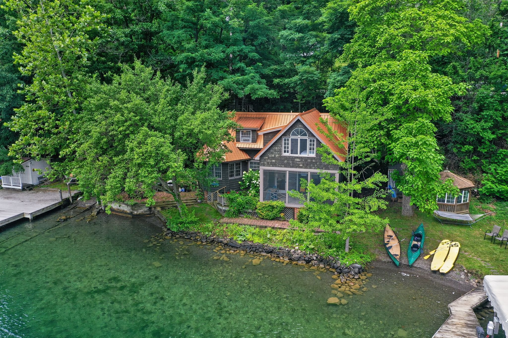 Una casa rodeada de árboles y una masa de agua, con un muelle y kayaks a la vista.