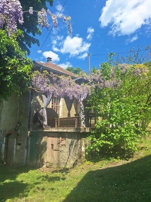 A house with a balcony, surrounded by greenery and purple flowers.
