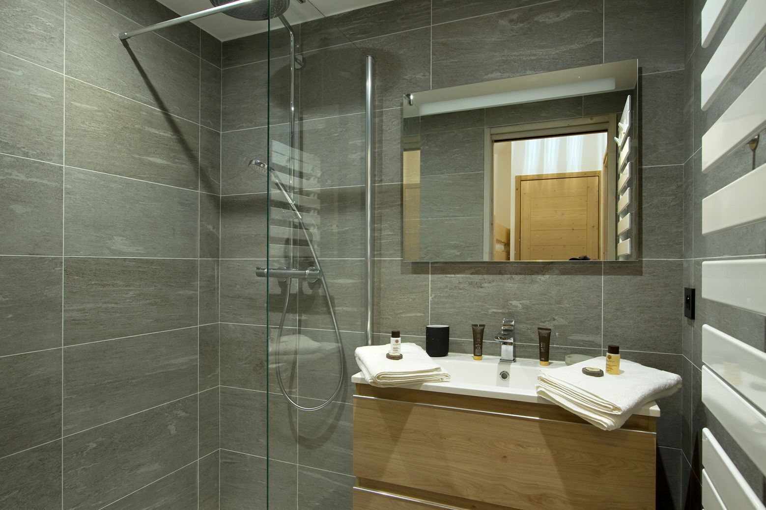Un baño moderno con ducha de vidrio, tocador de madera con espejo y suelo de baldosas.