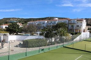 A tennis court with a net, surrounded by a green fence and a white building with multiple balconies. - Cvl Club House (Lagos)