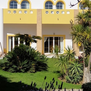 A building with a yellow facade, large windows, and a well-maintained garden with various plants. - Cvl Club House (Lagos)