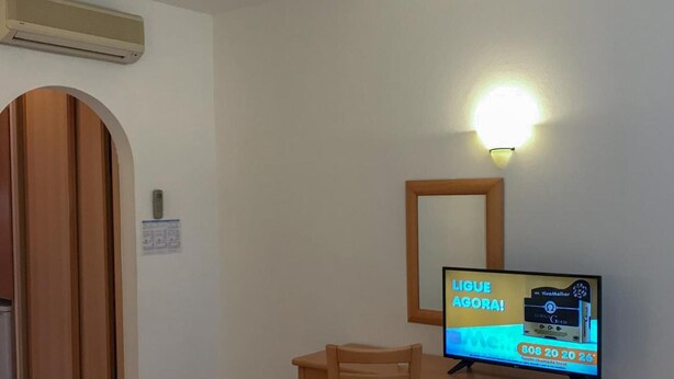 A hotel room with a wooden desk, a chair, a television displaying an advertisement, a wall-mounted mirror, and a door with a wooden frame. - Cvl Club House (Lagos)