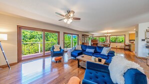 Un salon spacieux avec un canapé bleu, une table basse et un ventilateur de plafond. De grandes fenêtres offrent une vue sur la verdure environnante.