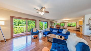 Un salon spacieux avec un canapé bleu, une table basse et un ventilateur de plafond. De grandes fenêtres offrent une vue sur la verdure environnante.