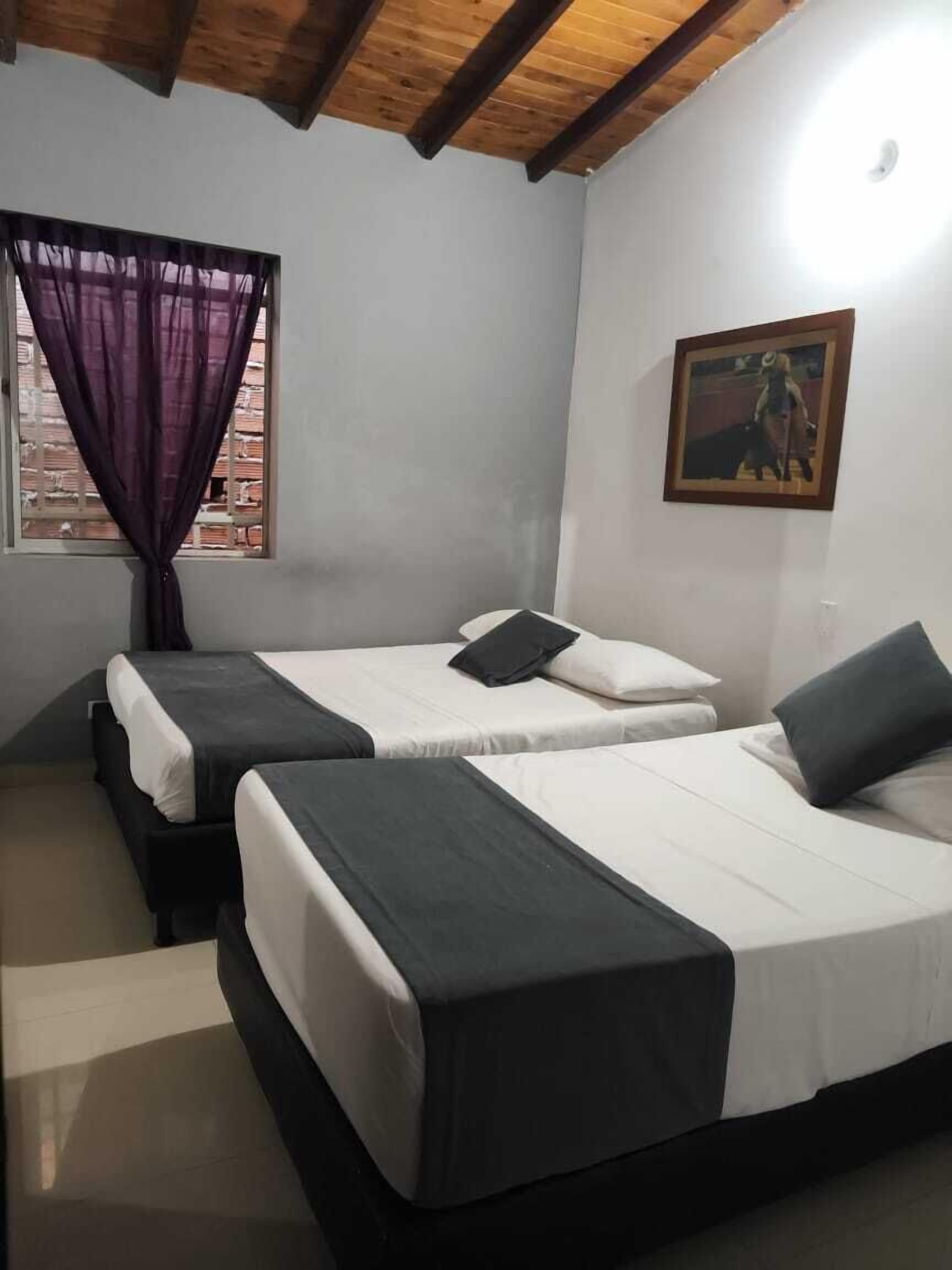 A room with two beds, a window with purple curtains, a painting on the wall, and a wooden ceiling.