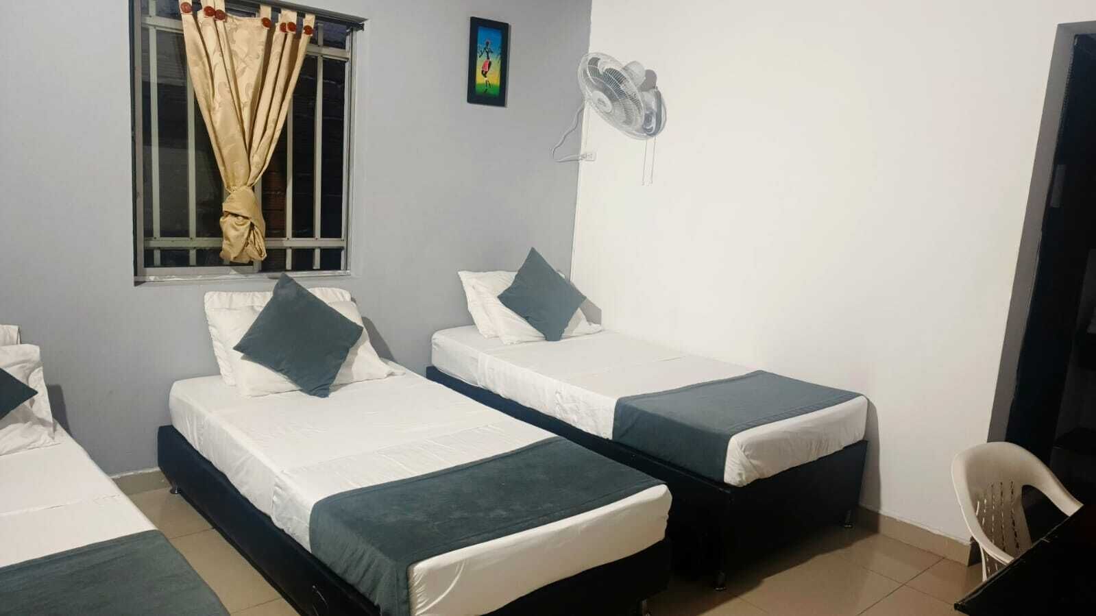 Une chambre avec deux lits simples, une fenêtre avec des rideaux, un ventilateur fixé au mur et une petite chaise.