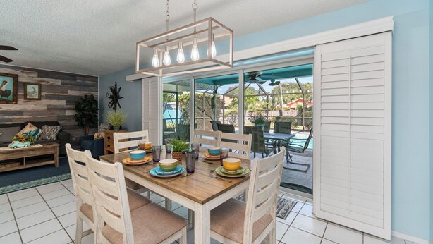 A dining area with a wooden table set for four, surrounded by white chairs. There is a ceiling fan, a hanging light fixture, and a view of the outdoors through a glass door.
