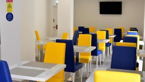 A modern dining area with yellow and blue chairs and white tables, a flat-screen TV on the wall, and a fire exit sign on the left.
