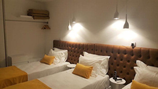 A hotel room with two single beds, each with white bedding and a yellow pillow. There are two pendant lights above the beds, and a small round table with a phone and a remote control.