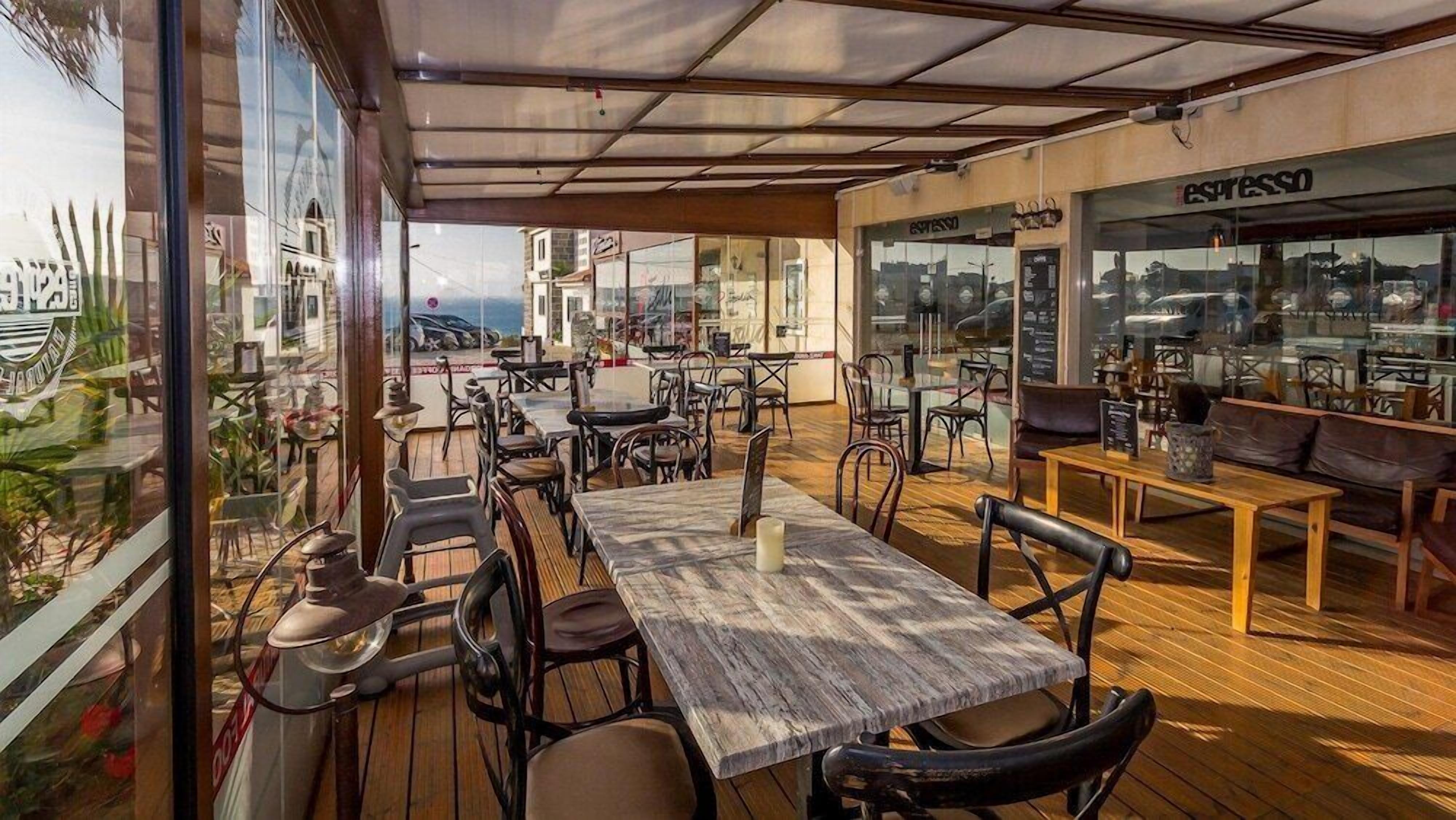 An outdoor café with wooden tables and chairs, a glass wall offering a view of the sea and buildings, and an espresso bar on the right.