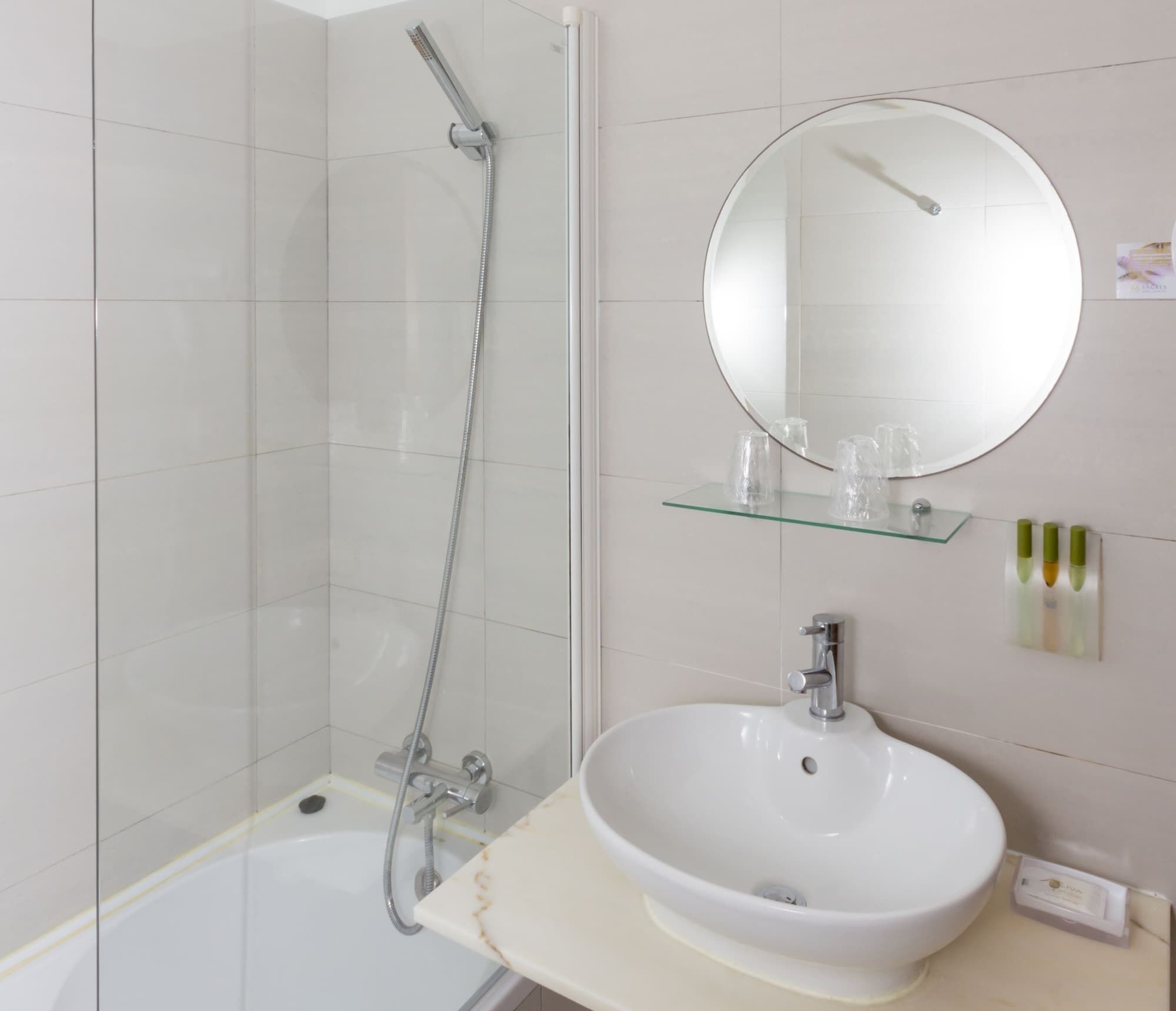 A bathroom with a glass shower enclosure, a white sink with a round mirror above it, and a glass shelf with toiletries.