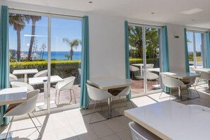 Una habitación de hotel moderna con grandes puertas de vidrio correderas que dan a un patio al aire libre con mesas y sillas blancas, y vistas al océano.