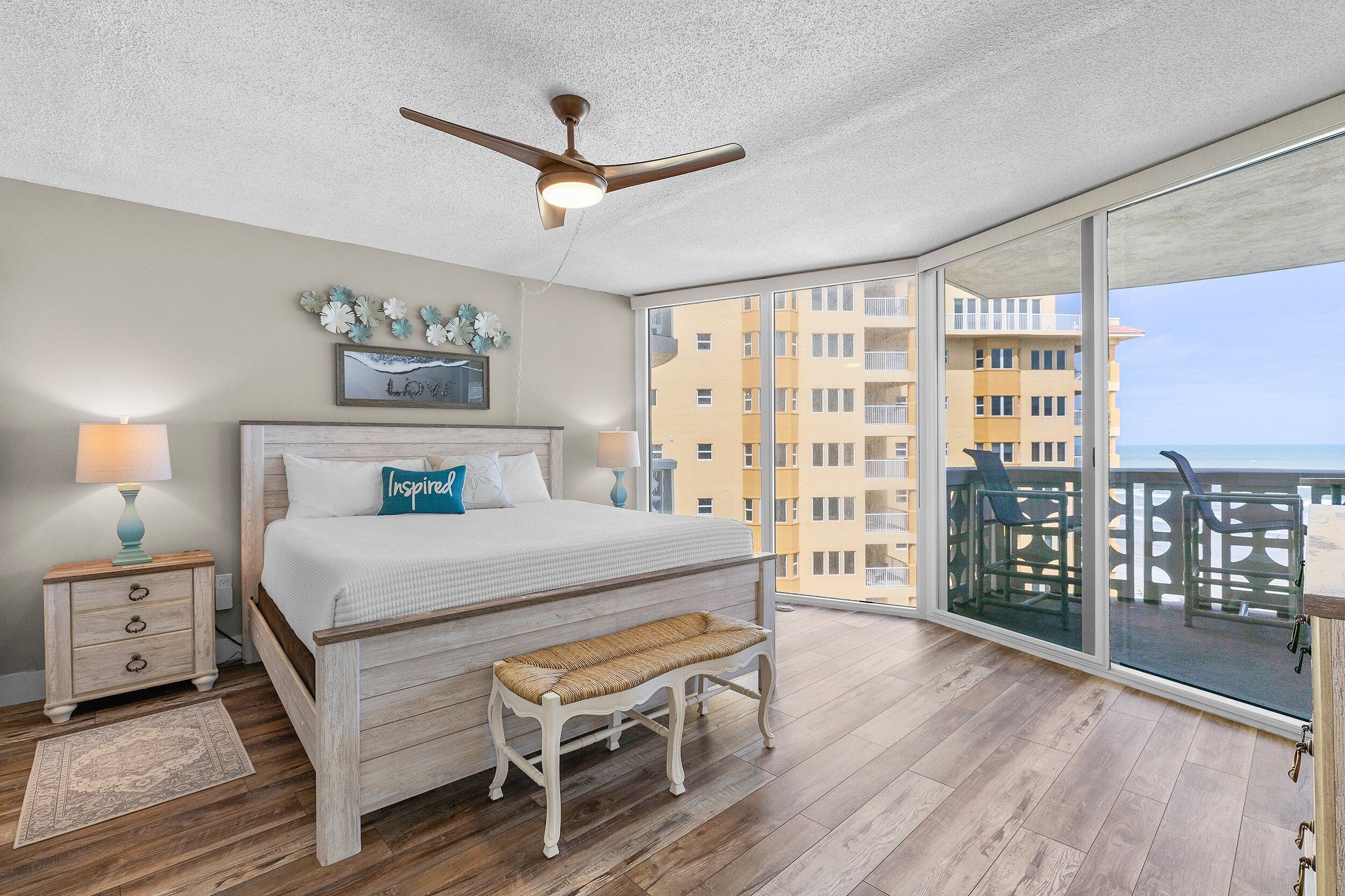 A bedroom with a large bed, a bench, a ceiling fan, and a balcony with a view of the city.