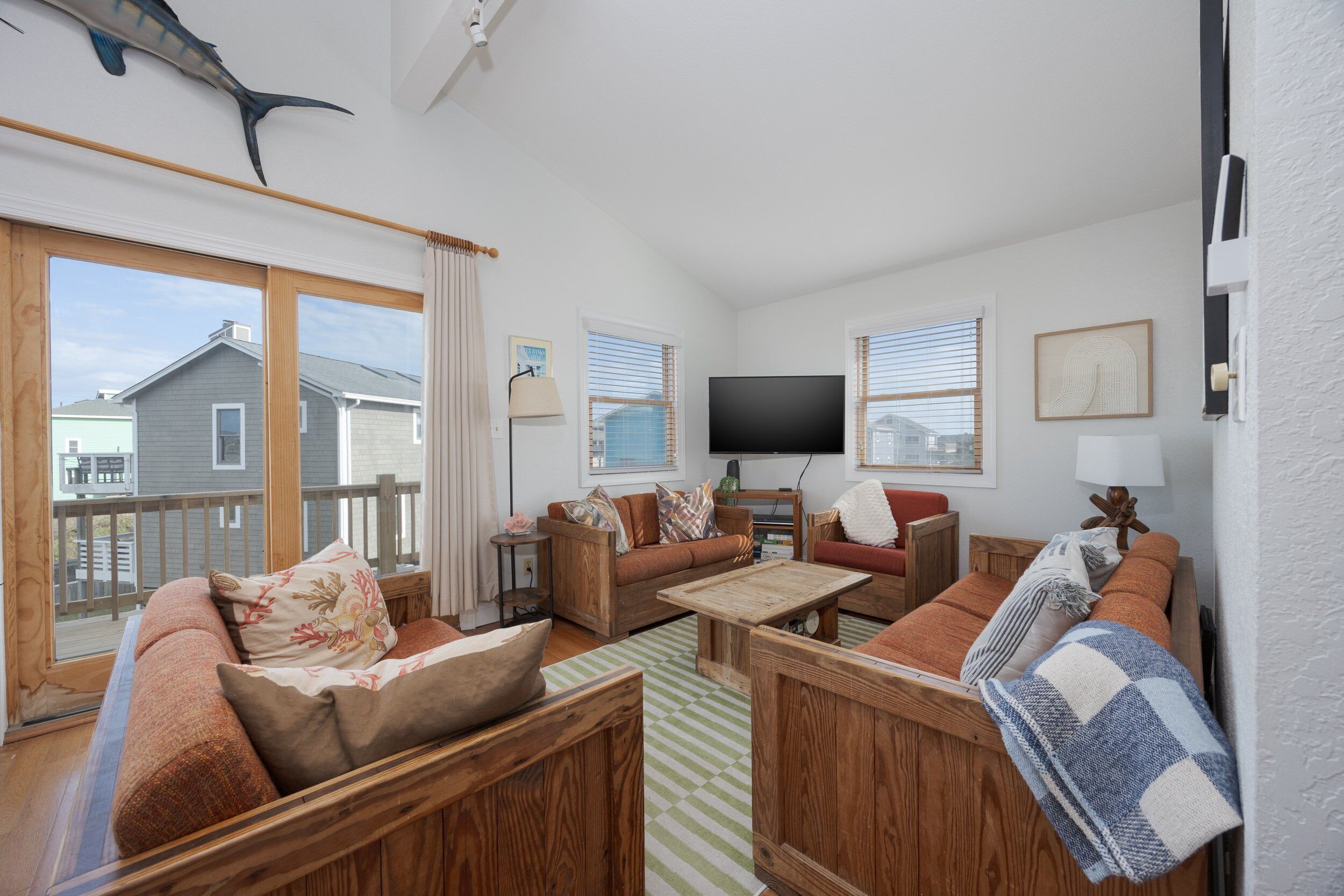 A cozy living room with wooden furniture, a flat-screen TV, and large windows with blinds.