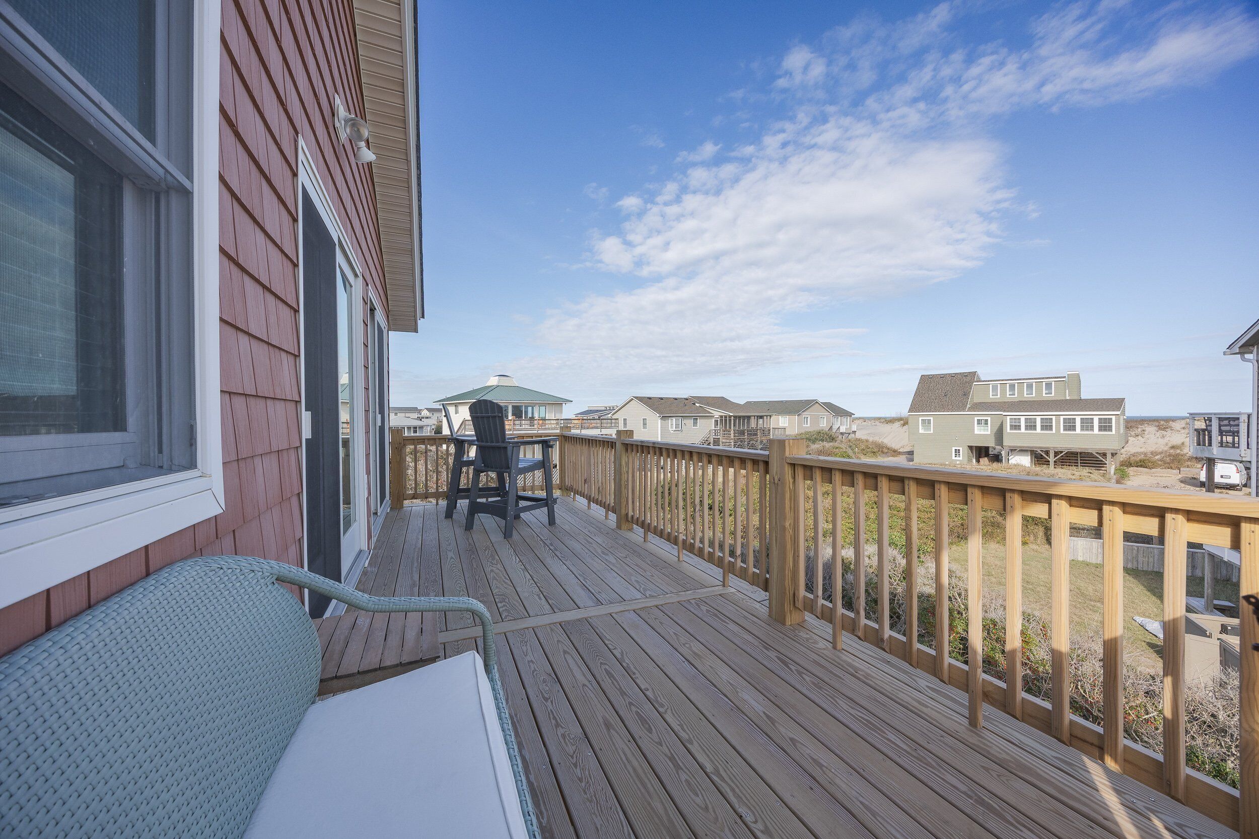 A wooden deck with a bench and a table, overlooking a beach with houses in the background.