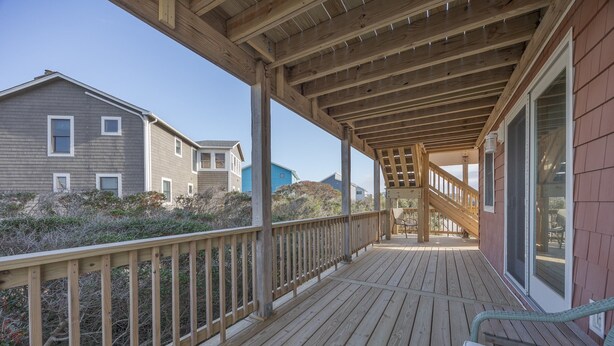 Una terraza de madera con barandilla, una escalera que lleva a un espacio cubierto y vista de casas vecinas.