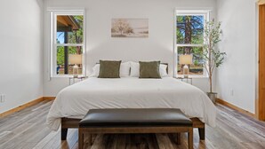 A bedroom with a large bed, two nightstands, a bench, and two windows with views of greenery.