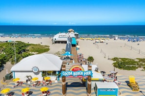 A beach pier with a sign reading "Cocoa Beach Pier" and "Westgate Resorts welcomes you to the beach." There are beach chairs and umbrellas, and the ocean is visible in the background.