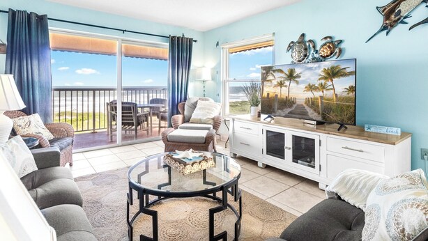 A living room with a view of the beach, a balcony with outdoor furniture, and a television on a white cabinet. - Gorgeous Direct Oceanfront w/ Private Balcony, Heated Pool & BBQ Grill! 🌊 (Cocoa Beach)