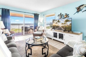 A living room with a view of the beach, a balcony with outdoor furniture, and a television on a white cabinet. - Gorgeous Direct Oceanfront w/ Private Balcony, Heated Pool & BBQ Grill! 🌊 (Cocoa Beach)