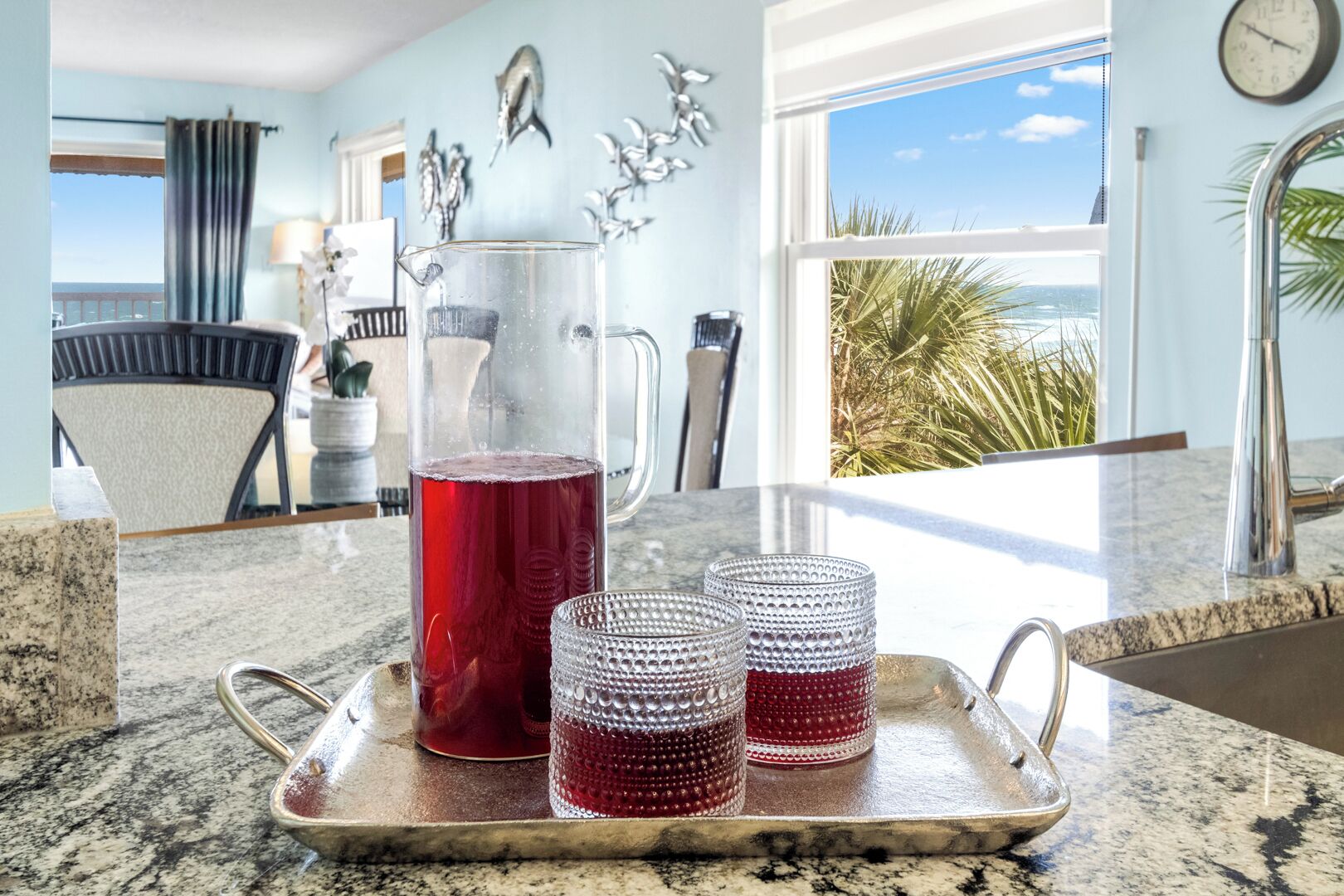 Una encimera de cocina con una jarra de bebida roja, dos vasos decorativos y una bandeja.