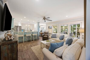 A spacious living room with a white sofa, a coffee table, and a TV. There is a kitchen area with a refrigerator and a dining table. - Tennis View! Updated Villa! WALK TO TENNIS CENTER! Screened Porch! (Kiawah Island)