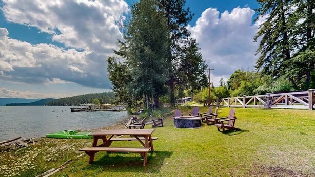 Ein Picknickplatz am Seeufer mit Holzbänken und -stühlen, einer Feuerstelle und Blick auf den See sowie ferne Berge.