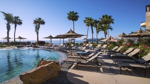 Un espace aménagé au bord de la piscine, avec des chaises longues et des parasols, des palmiers et une vue sur l’océan.