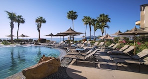Un espace aménagé au bord de la piscine, avec des chaises longues et des parasols, des palmiers et une vue sur l’océan.