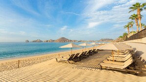 Une plage bordée de rangées de chaises longues et de parasols, de palmiers, et offrant une vue dégagée sur l’océan et les montagnes lointaines.